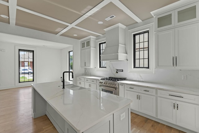 kitchen with premium range hood, light stone counters, high end stove, a kitchen island with sink, and white cabinetry