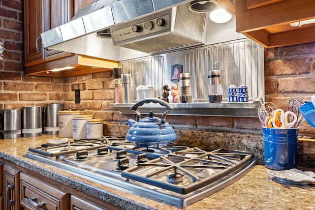 kitchen with stainless steel gas stovetop, stone counters, exhaust hood, and brick wall