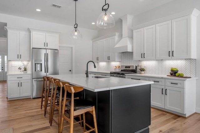 kitchen with white cabinets, appliances with stainless steel finishes, wall chimney exhaust hood, an island with sink, and hanging light fixtures