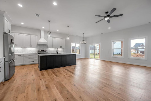 kitchen featuring white cabinetry, a center island with sink, stainless steel appliances, decorative light fixtures, and wall chimney exhaust hood