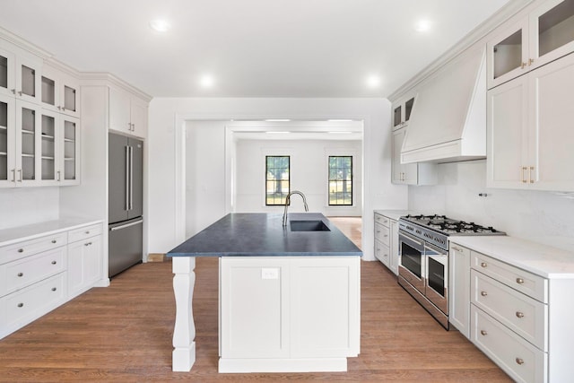 kitchen with premium appliances, white cabinets, custom exhaust hood, sink, and a kitchen island with sink