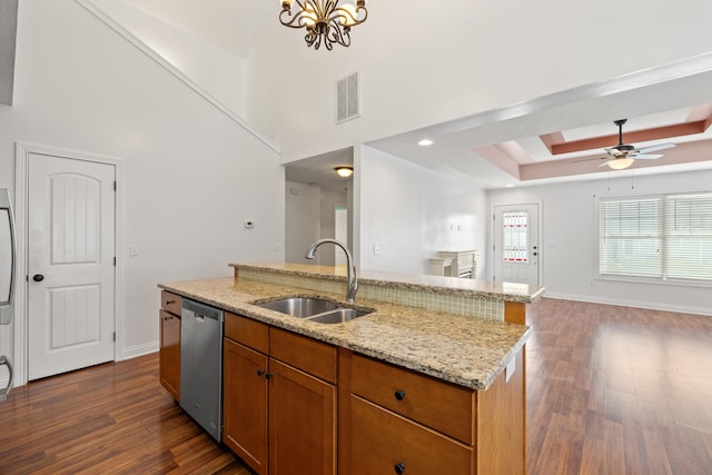 kitchen featuring stainless steel dishwasher, sink, a tray ceiling, light stone countertops, and a center island with sink