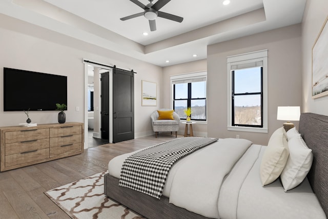 bedroom with hardwood / wood-style floors, ensuite bath, a barn door, ceiling fan, and a tray ceiling