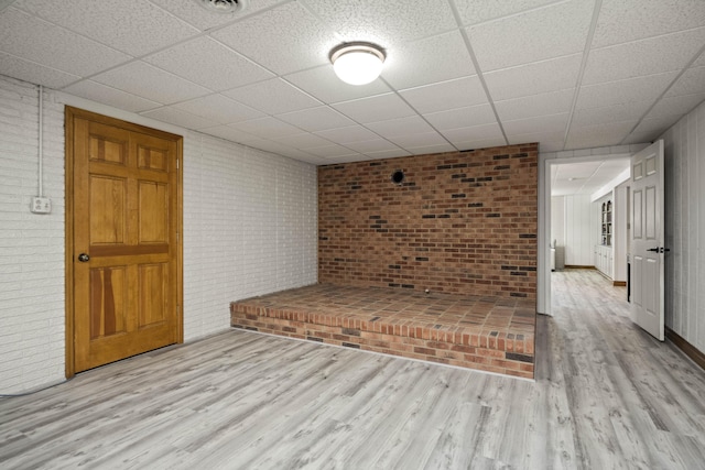 unfurnished living room featuring brick wall, a drop ceiling, and light hardwood / wood-style flooring