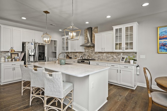 kitchen featuring wall chimney exhaust hood, appliances with stainless steel finishes, pendant lighting, a kitchen island with sink, and white cabinets