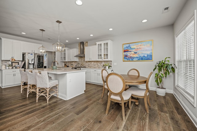 kitchen with white cabinetry, hanging light fixtures, wall chimney range hood, stainless steel appliances, and a center island with sink