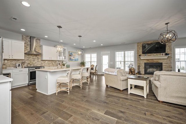 kitchen with stainless steel stove, decorative light fixtures, an island with sink, white cabinets, and wall chimney range hood