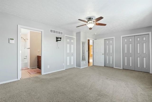 unfurnished bedroom featuring ensuite bath, ceiling fan, a textured ceiling, two closets, and light colored carpet