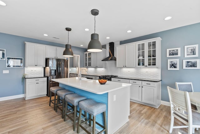 kitchen with pendant lighting, an island with sink, stainless steel fridge, wall chimney range hood, and light hardwood / wood-style flooring