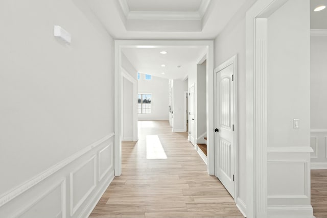 corridor featuring light wood finished floors, a raised ceiling, a wainscoted wall, crown molding, and a decorative wall