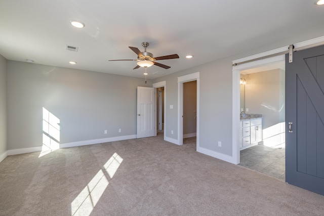 unfurnished bedroom featuring ceiling fan, light colored carpet, a barn door, and ensuite bath