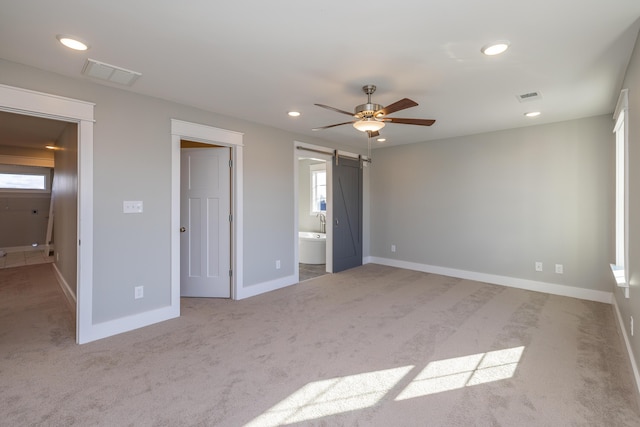 unfurnished bedroom featuring ensuite bath, light colored carpet, a barn door, and ceiling fan