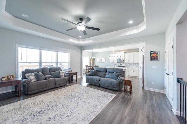 living area with dark wood finished floors, recessed lighting, a raised ceiling, baseboards, and ceiling fan with notable chandelier