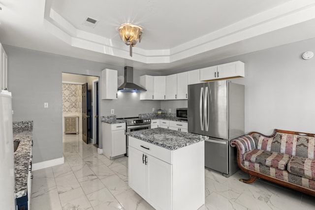 kitchen featuring a kitchen island, a raised ceiling, white cabinetry, stainless steel appliances, and wall chimney exhaust hood