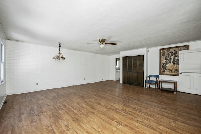 unfurnished living room featuring dark wood-style floors, brick wall, and ceiling fan with notable chandelier