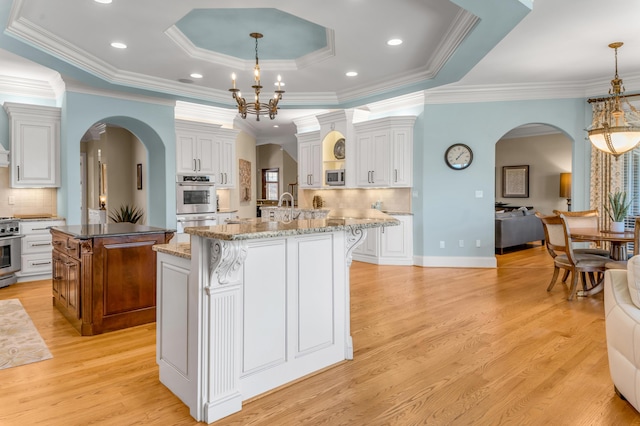 kitchen featuring a raised ceiling, a kitchen island with sink, hanging light fixtures, and white cabinets