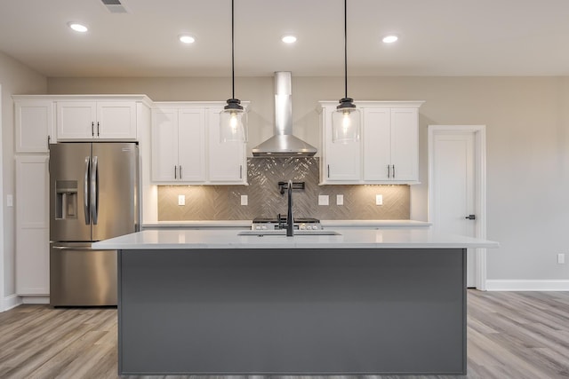 kitchen with light countertops, wall chimney range hood, an island with sink, and stainless steel fridge