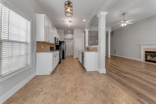 kitchen with ceiling fan, stainless steel appliances, a tiled fireplace, decorative columns, and white cabinets