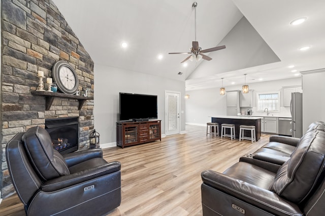 living area featuring light wood-style flooring, a ceiling fan, a stone fireplace, high vaulted ceiling, and baseboards