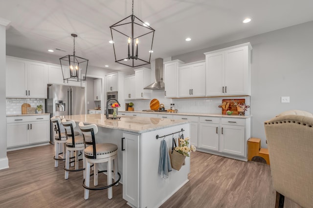 kitchen with white cabinets, a center island with sink, hanging light fixtures, and wall chimney range hood