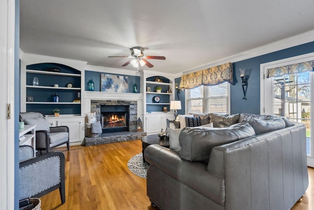 living room with built in features, ceiling fan, a fireplace, ornamental molding, and light wood-type flooring