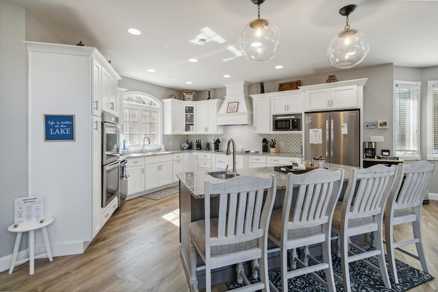 kitchen featuring premium range hood, decorative light fixtures, an island with sink, stainless steel appliances, and light stone countertops