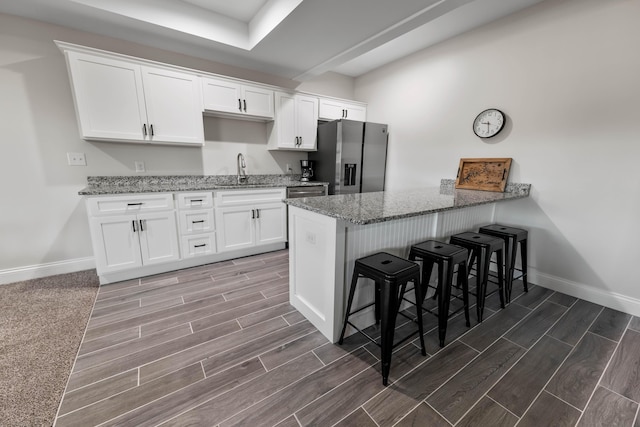 kitchen featuring stainless steel refrigerator with ice dispenser, a kitchen bar, sink, white cabinetry, and light stone counters