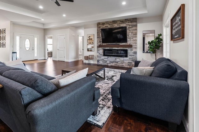 living room featuring a tray ceiling, dark wood-type flooring, a stone fireplace, and ceiling fan