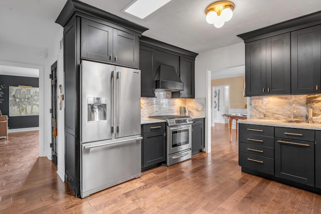 kitchen featuring appliances with stainless steel finishes, hardwood / wood-style floors, light countertops, wall chimney range hood, and backsplash