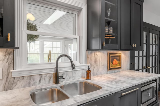 kitchen featuring light stone counters, a skylight, a sink, stainless steel microwave, and glass insert cabinets