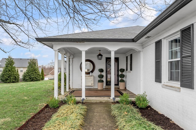 doorway to property with a shingled roof, covered porch, brick siding, and a yard