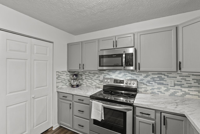 kitchen with gray cabinets, appliances with stainless steel finishes, backsplash, dark hardwood / wood-style flooring, and light stone counters