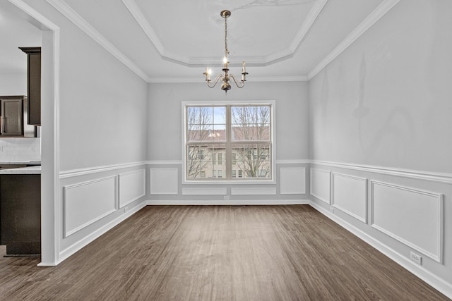 unfurnished dining area featuring a decorative wall, dark wood-style floors, an inviting chandelier, a raised ceiling, and crown molding