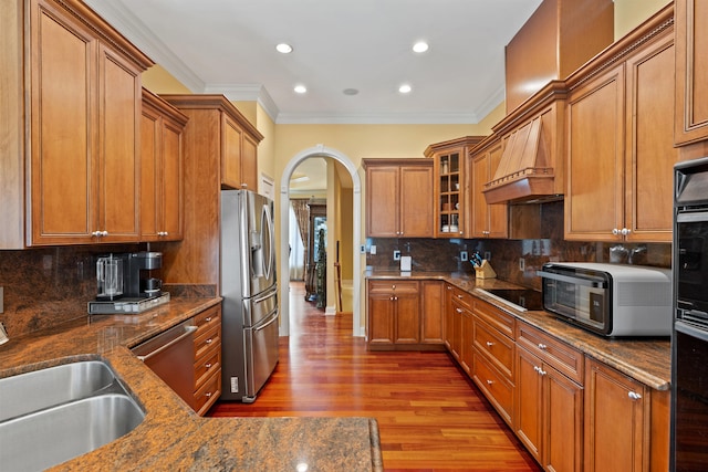 kitchen with arched walkways, stainless steel appliances, brown cabinetry, wood finished floors, and dark stone counters