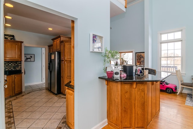 kitchen with brown cabinets, stainless steel refrigerator with ice dispenser, dark stone countertops, a peninsula, and baseboards