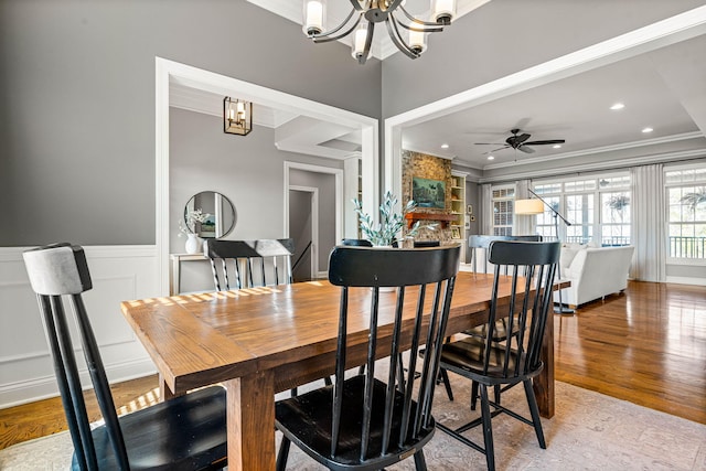 dining area featuring ornamental molding, a stone fireplace, ceiling fan with notable chandelier, and light wood-type flooring