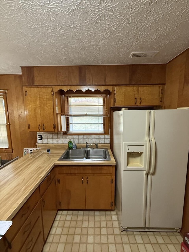 kitchen featuring sink, backsplash, wooden walls, white refrigerator with ice dispenser, and a textured ceiling