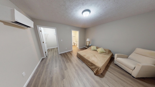 bedroom featuring a wall mounted air conditioner, a spacious closet, a textured ceiling, ensuite bath, and light hardwood / wood-style flooring