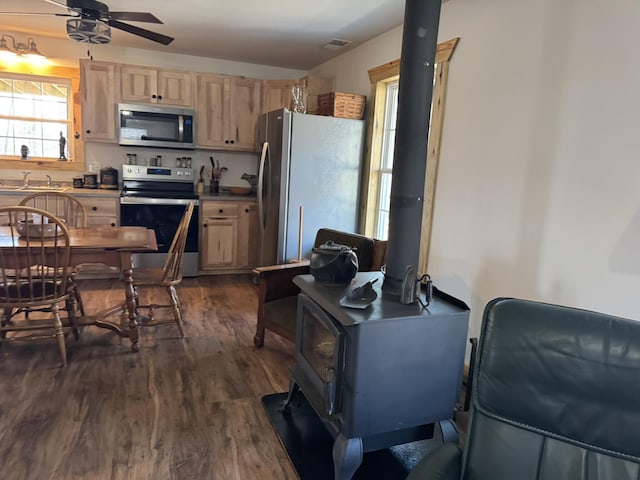 kitchen with dark wood-type flooring, ceiling fan, appliances with stainless steel finishes, light brown cabinets, and a wood stove