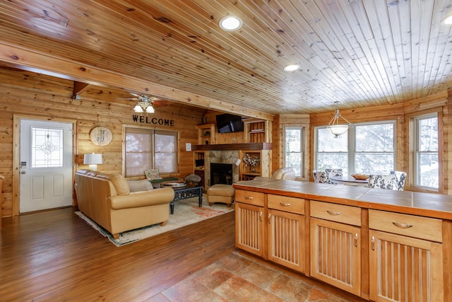 kitchen with a stone fireplace, wooden walls, tile counters, pendant lighting, and a healthy amount of sunlight