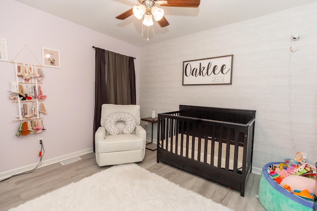 bedroom with visible vents, light wood-style flooring, ceiling fan, a nursery area, and baseboards