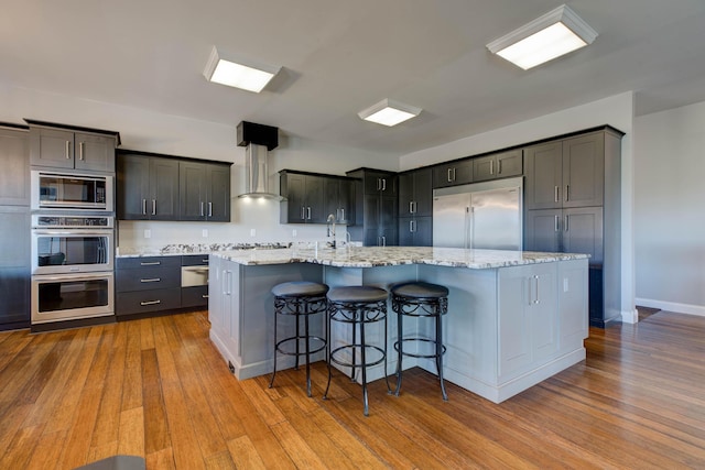 kitchen featuring wood finished floors, a spacious island, a breakfast bar, built in appliances, and wall chimney exhaust hood