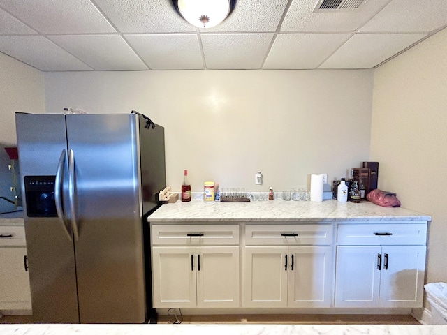 kitchen featuring stainless steel refrigerator with ice dispenser, white cabinetry, light stone countertops, and a drop ceiling