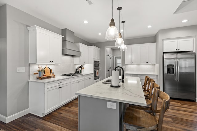 kitchen featuring appliances with stainless steel finishes, a kitchen island with sink, premium range hood, white cabinets, and hanging light fixtures