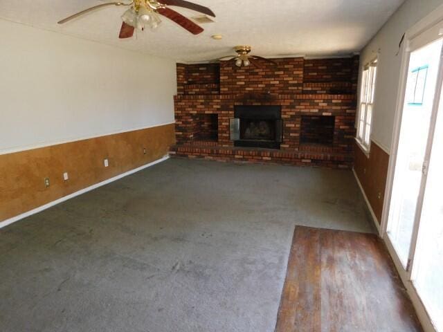 unfurnished living room featuring ceiling fan, wooden walls, wainscoting, a brick fireplace, and dark carpet