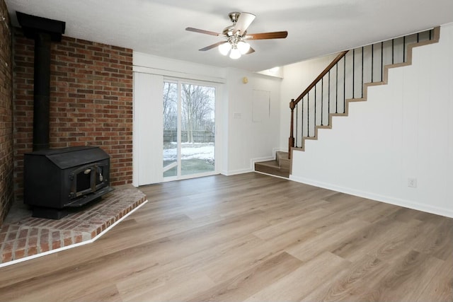 unfurnished living room featuring baseboards, light wood-style flooring, ceiling fan, stairway, and a wood stove