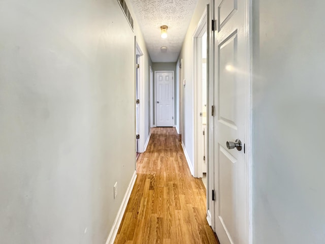 hall with light wood-type flooring, visible vents, a textured ceiling, and baseboards