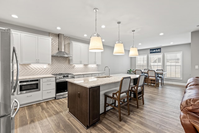 kitchen with a kitchen island with sink, stainless steel appliances, a sink, white cabinets, and wall chimney exhaust hood