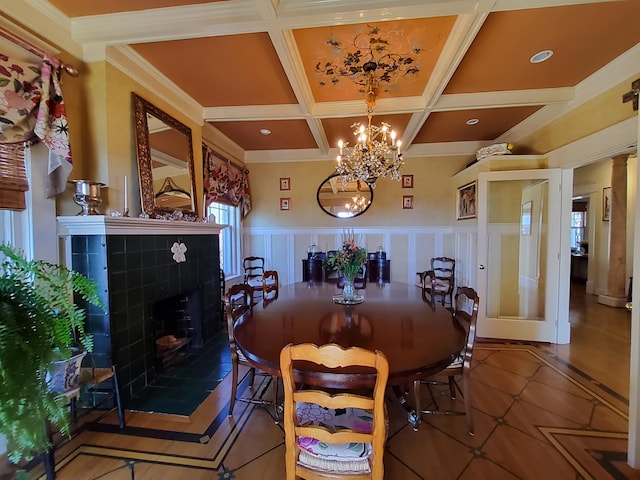 dining space with a chandelier, a wainscoted wall, coffered ceiling, a tiled fireplace, and crown molding