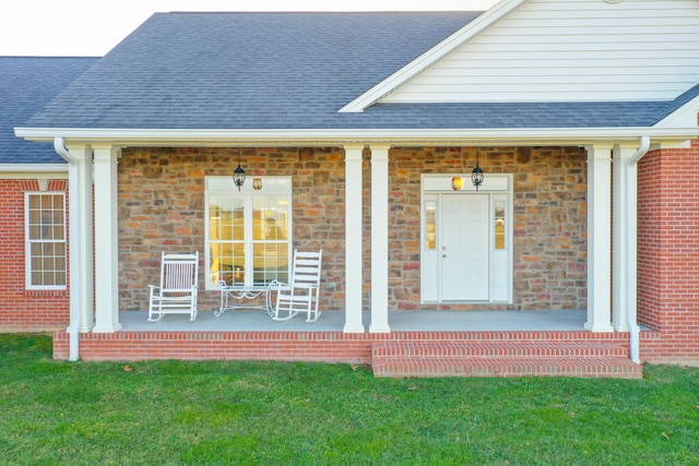 doorway to property featuring a yard, a porch, a shingled roof, and brick siding